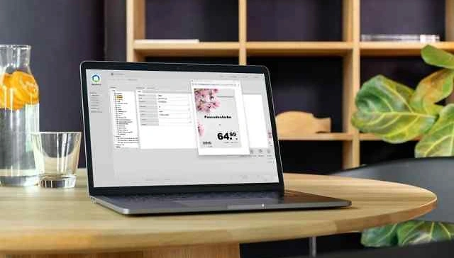 Laptop auf rundem Holztisch mit ge&ouml;ffneter Software f&uuml;r Plakatdruck, daneben Glas mit Wasser und Zitronenscheiben, im Hintergrund Regal und Pflanze