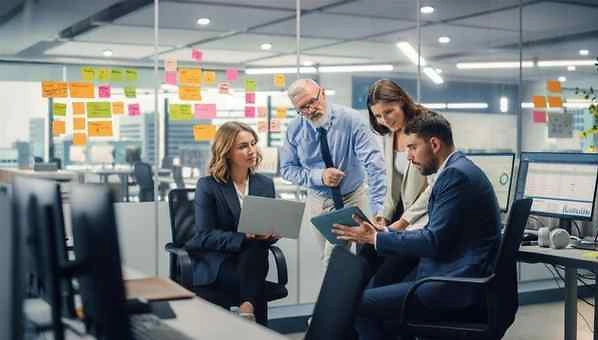 Vier Personen in einem modernen B&uuml;ro besprechen Dokumente und digitale Ger&auml;te vor einer Glaswand mit vielen bunten Haftnotizen