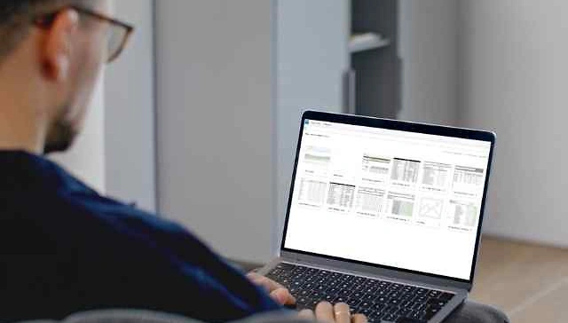 Person mit Brille sitzt an einem Tisch und arbeitet an einem Laptop mit ge&ouml;ffnetem Bildschirm, der mehrere Tabellen zeigt