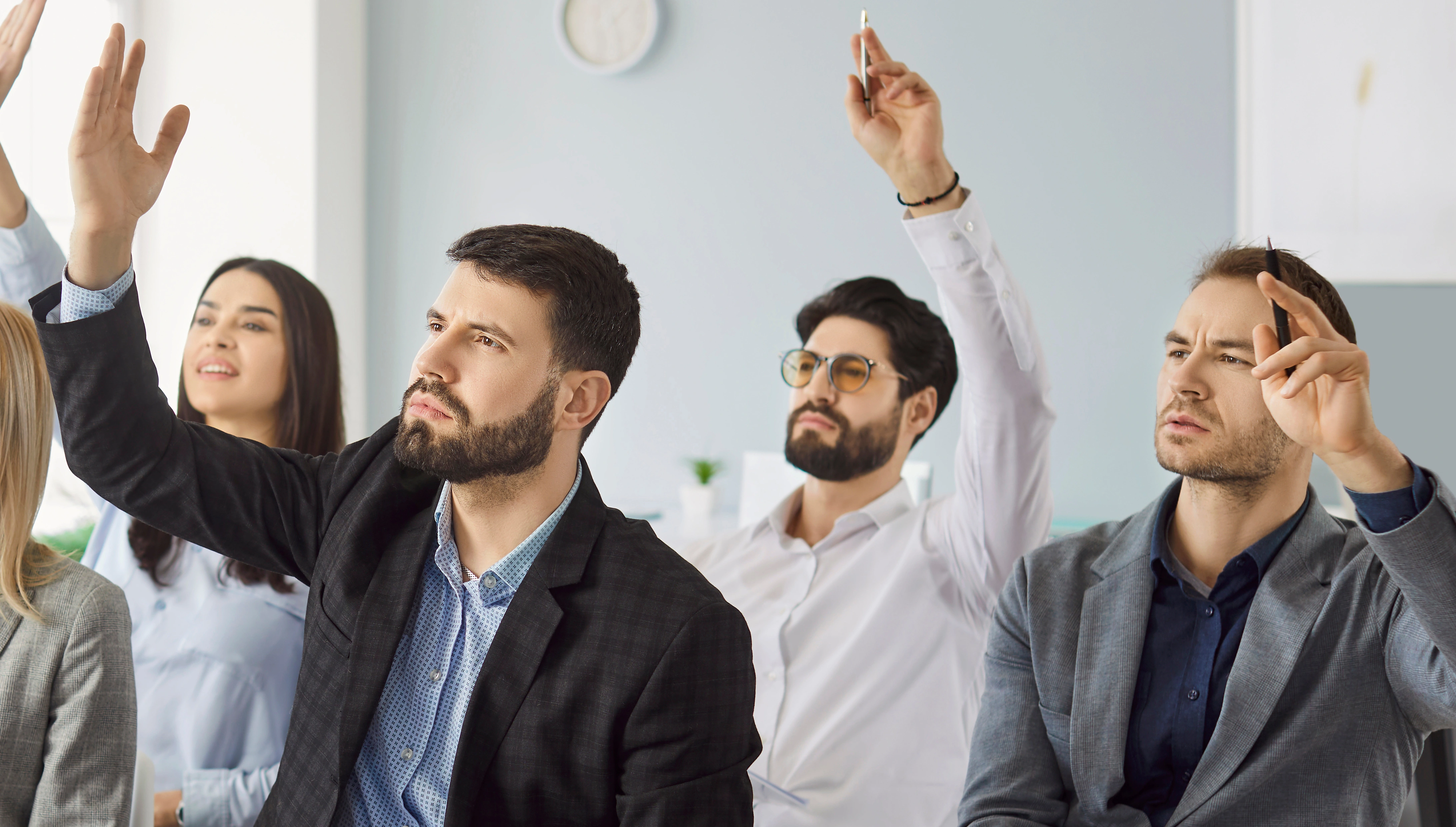 F&uuml;nf Personen in Businesskleidung sitzen in einem Raum und heben jeweils eine Hand